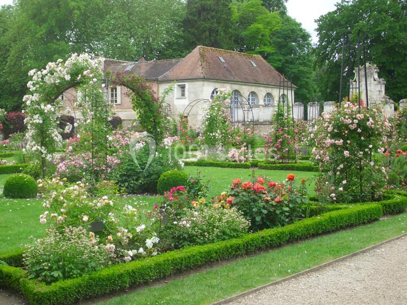 Jardin fleuri avec rosiers, allées gravillonnées et petite maison en arrière-plan entourée de verdure.