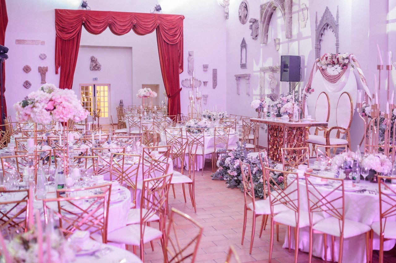 Salle de réception décorée pour un mariage avec des tables ornées de fleurs roses et blanches, chaises dorées et arche…