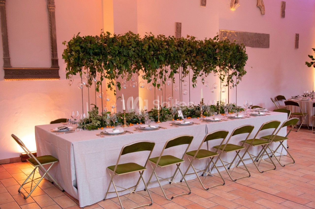 Table de réception décorée avec des chandeliers, feuillage suspendu et vaisselle élégante dans une salle lumineuse.