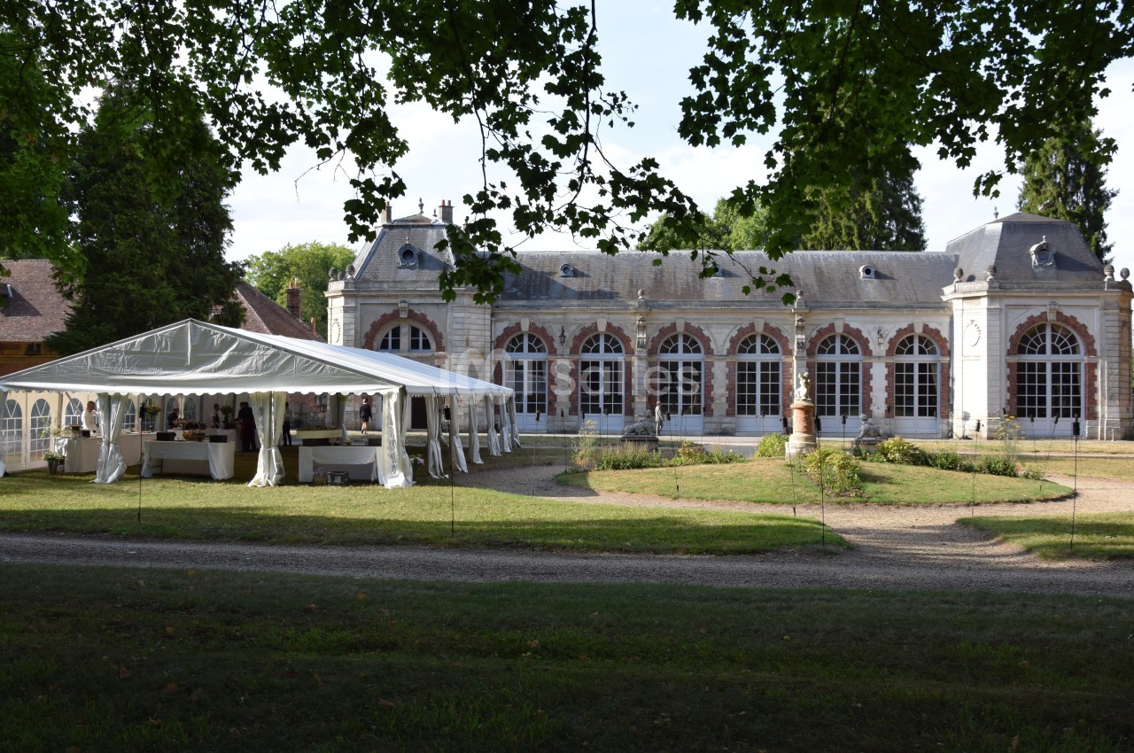Bâtiment ancien avec de grandes fenêtres en arc, entouré d'arbres, et une tente blanche installée sur la pelouse.