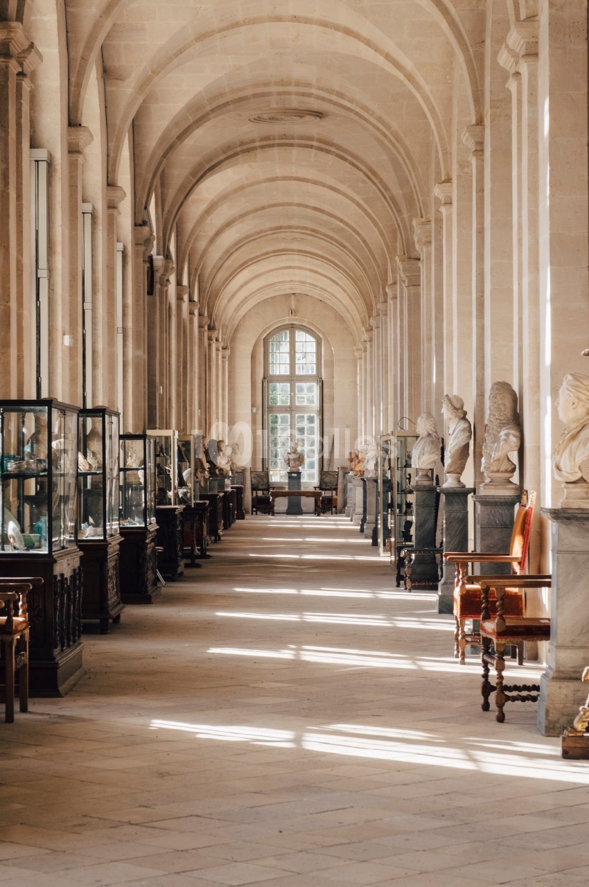 Galerie lumineuse avec des vitrines, des sculptures et des chaises, sous une voûte en pierre avec des arcs en enfilade.