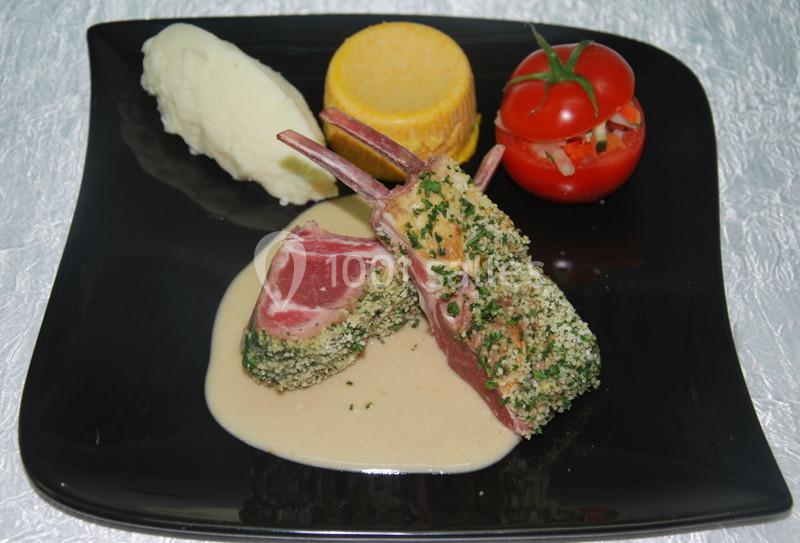 Carré d'agneau en croûte d'herbes sur sauce, accompagné de purée, flan de légumes et tomate farcie, servi sur assiette noire.