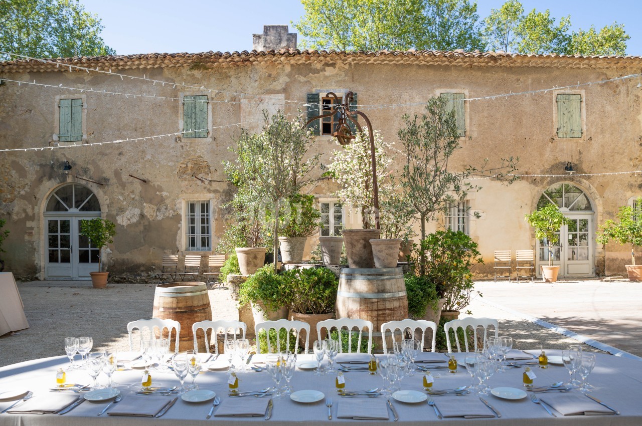 Table dressée en extérieur avec vaisselle élégante, devant une bâtisse ancienne entourée de verdure et de pots de fleurs.