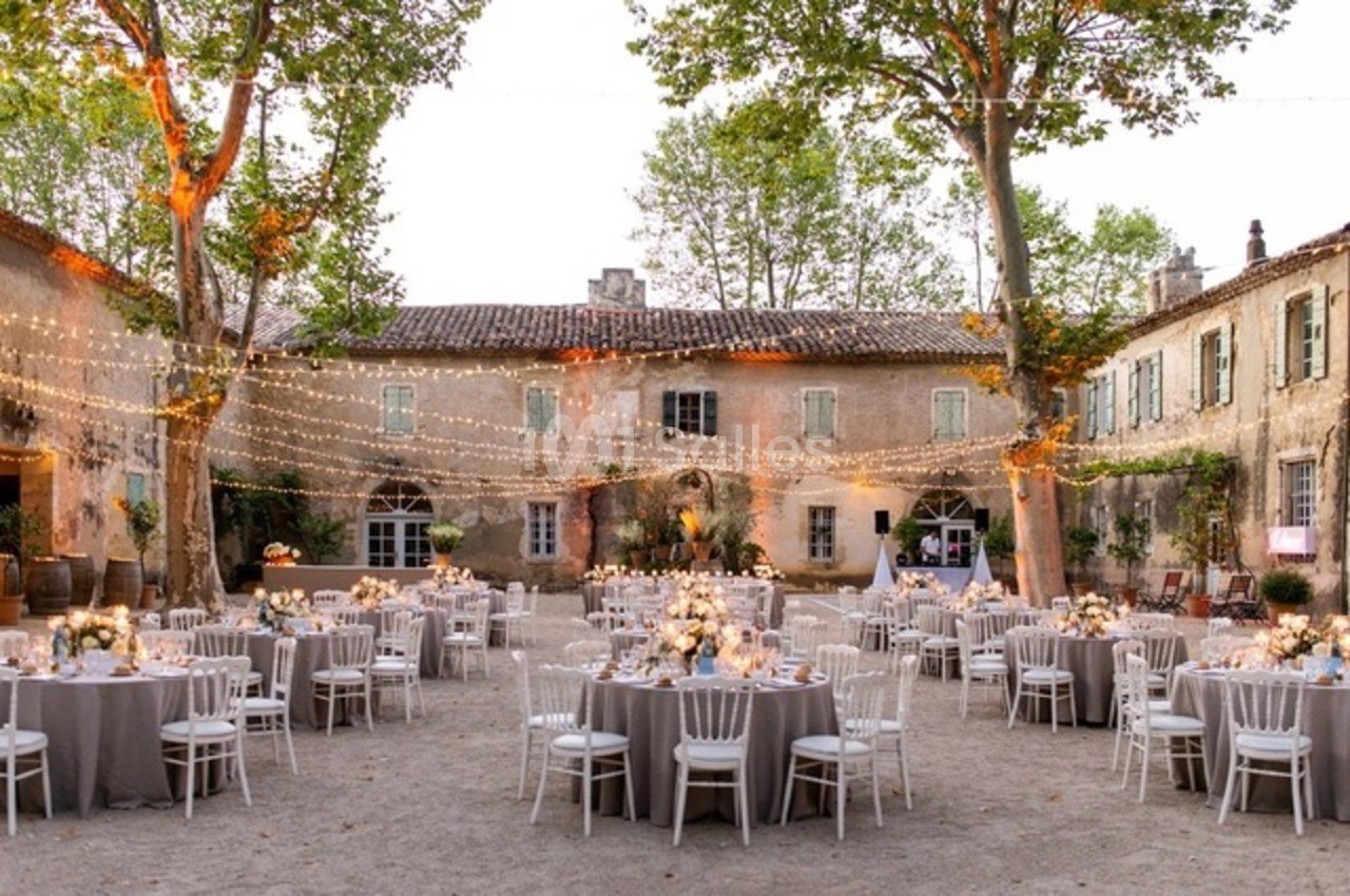 Cour d'un domaine ancien décorée pour un mariage, avec tables dressées, chaises blanches et guirlandes lumineuses suspendues.