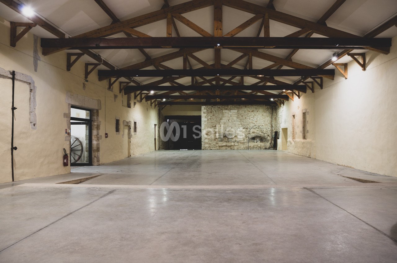 Intérieur d'une grande salle vide avec plafond en poutres apparentes et murs en pierre et plâtre.