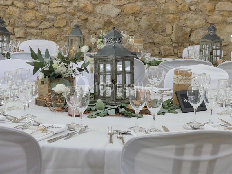 Table décorée pour un mariage avec lanternes, fleurs blanches, feuillage et vaisselle élégante, devant un mur en pierre.
