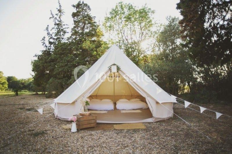 Tente en toile beige installée sur un sol gravillonné, entourée d'arbres, avec des lits aménagés à l'intérieur.