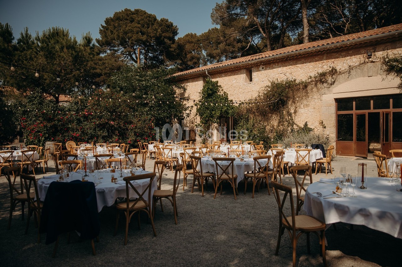 Tables rondes dressées avec nappes blanches et chaises en bois dans une cour extérieure entourée de végétation et de murs en…