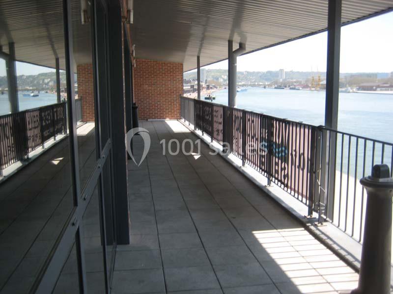 Balcon couvert avec sol carrelé, vue sur un fleuve et des bâtiments au loin, encadré par une rambarde métallique.