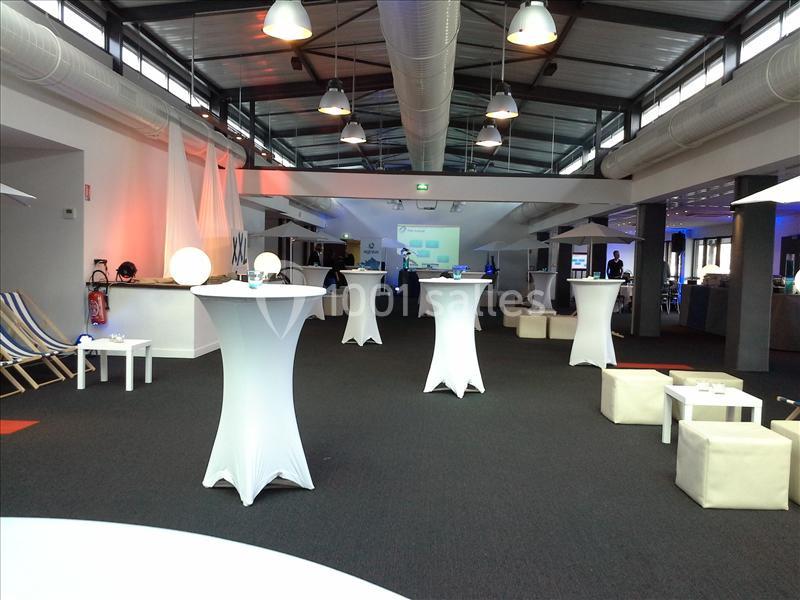 Salle aménagée pour un événement avec tables hautes drapées de blanc, poufs et éclairage tamisé.