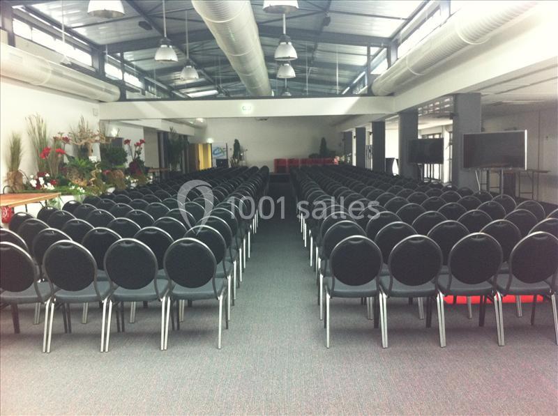Salle de conférence avec des rangées de chaises noires alignées sous un plafond industriel avec des conduits apparents.