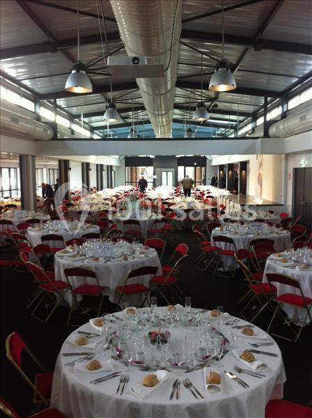Salle de réception avec tables rondes dressées, nappes blanches, chaises rouges et couverts disposés pour un événement.