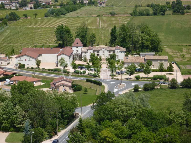 Vue aérienne d'un village avec des bâtiments, des routes, des arbres et des vignobles en arrière-plan.