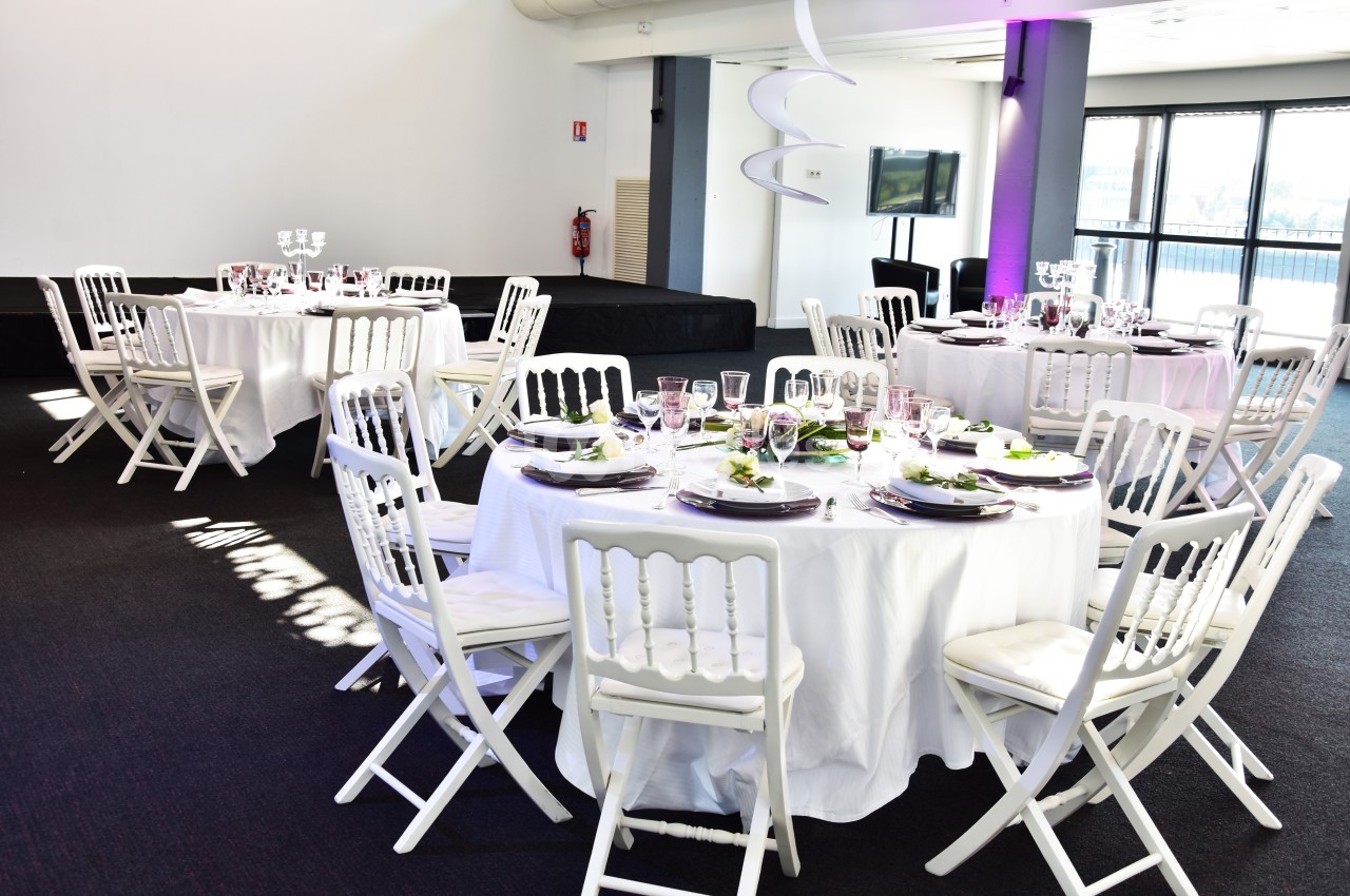 Salle lumineuse avec tables rondes dressées, nappes blanches, chaises pliantes blanches et décoration élégante.