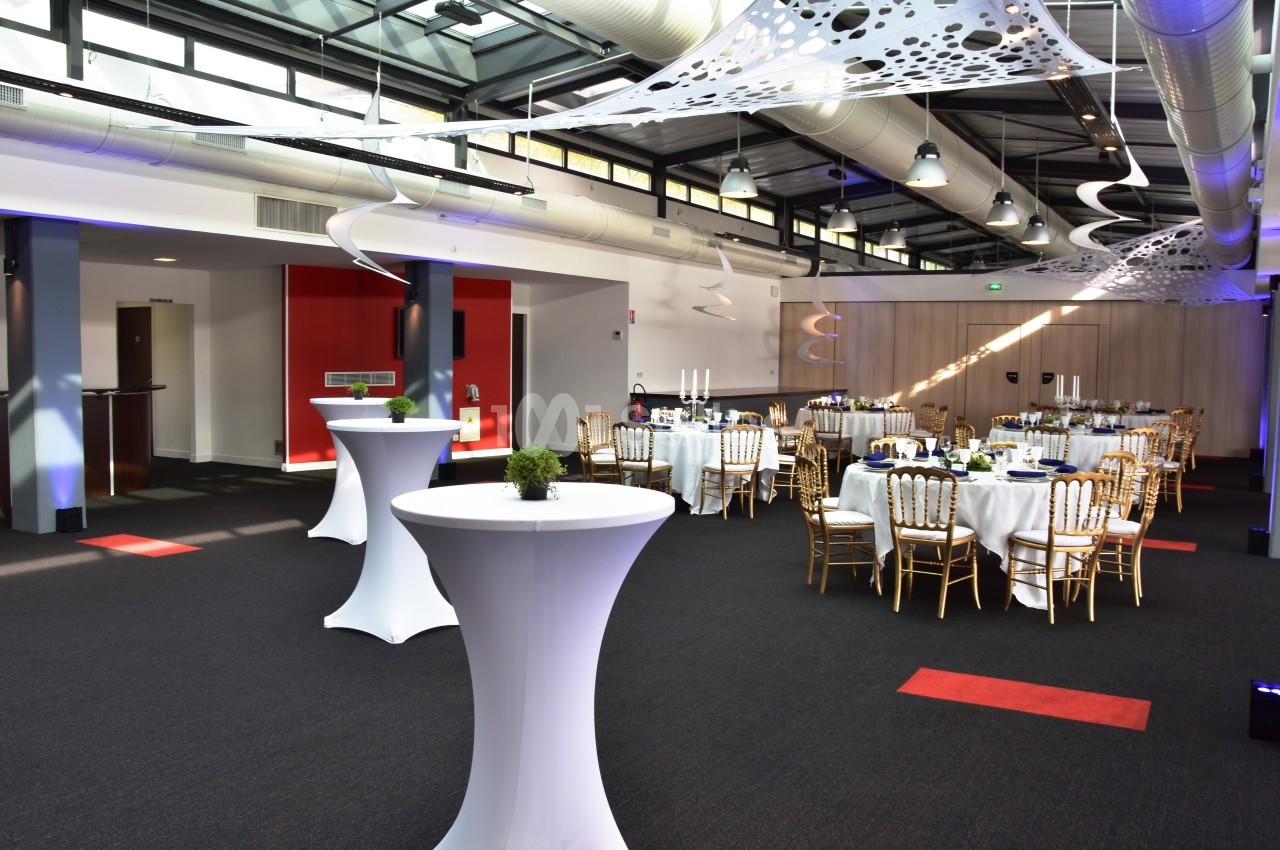 Salle de réception moderne avec tables rondes dressées, chaises dorées et décorations suspendues.