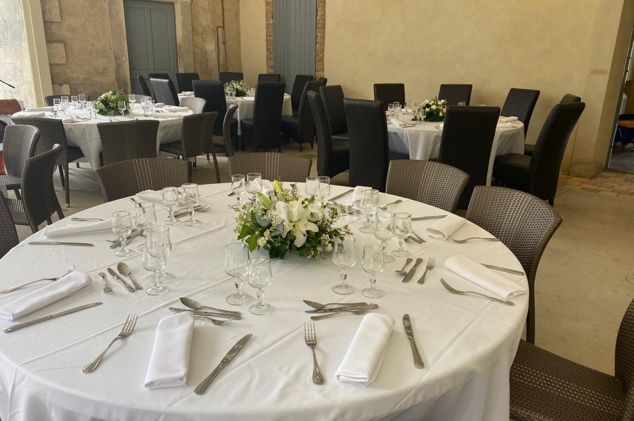 Salle de réception avec tables rondes dressées, nappes blanches, couverts, verres et centres de table fleuris.