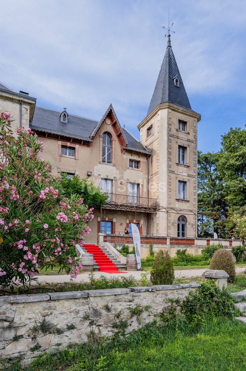 Façade d'un manoir avec une tour, escalier rouge, jardin fleuri et ciel dégagé en arrière-plan.
