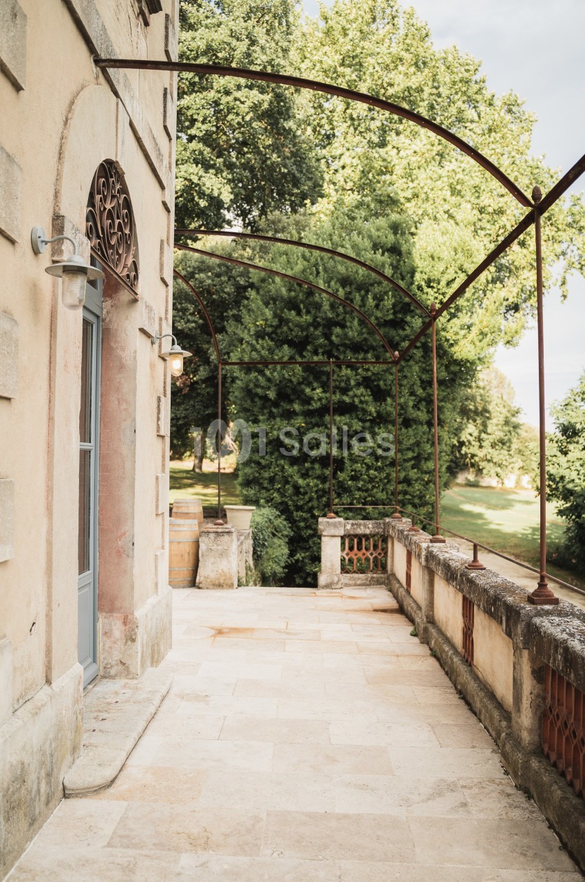 Terrasse en pierre bordée de balustrades et d'arches métalliques, donnant sur un jardin arboré.