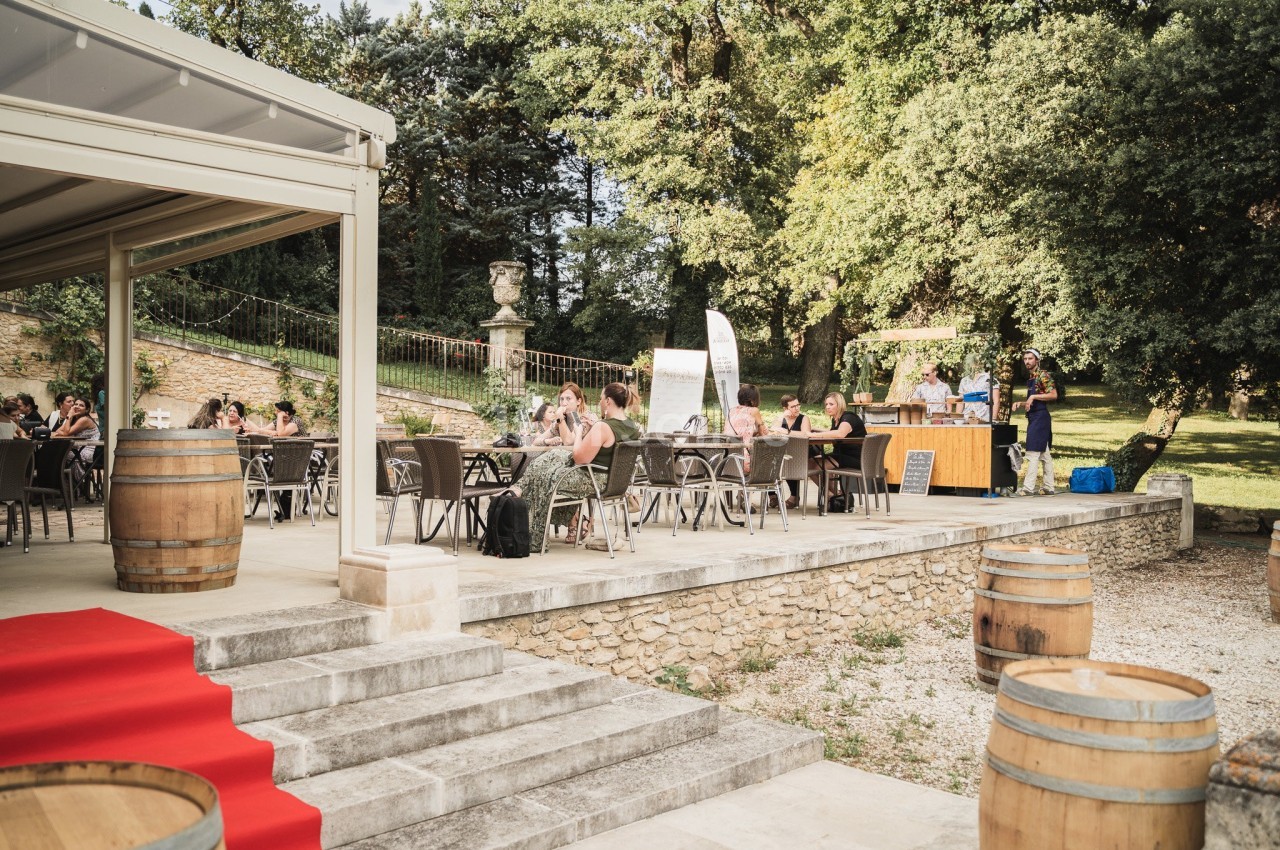 Personnes assises à des tables en extérieur près d'un bâtiment ouvert, entourées de verdure et de tonneaux en bois.