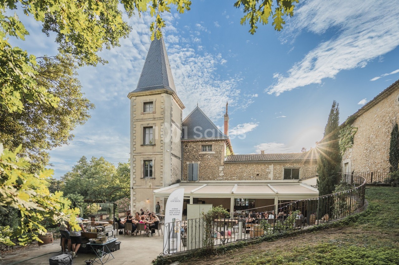 Vue d'un bâtiment en pierre avec une tour, entouré de verdure, abritant une terrasse animée par des convives.