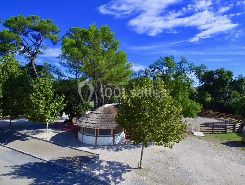 Abri circulaire au toit de chaume entouré d'arbres, avec une clôture en bois et un ciel bleu dégagé.