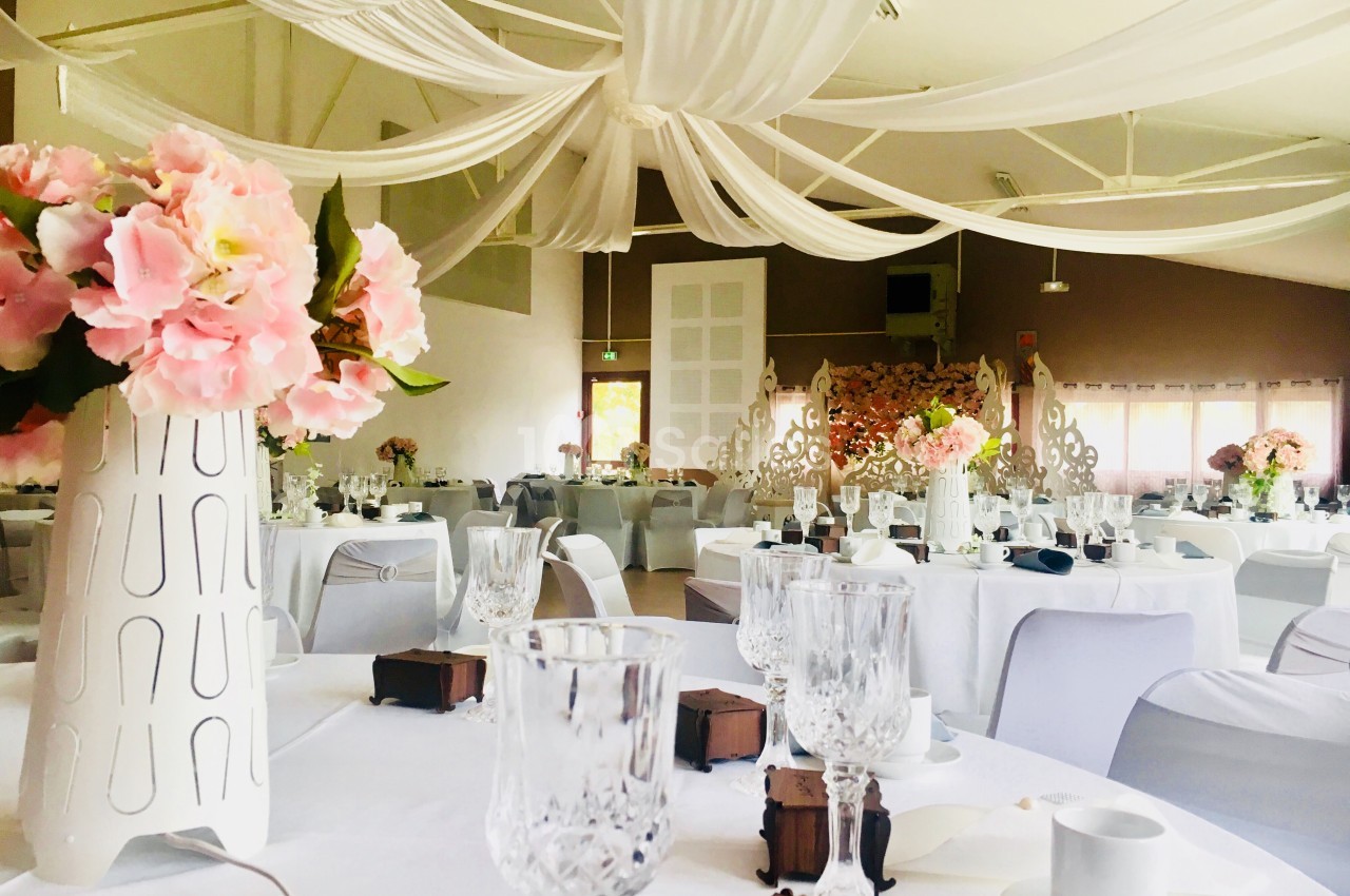 Salle de réception décorée avec des nappes blanches, des fleurs roses et des drapés suspendus au plafond.