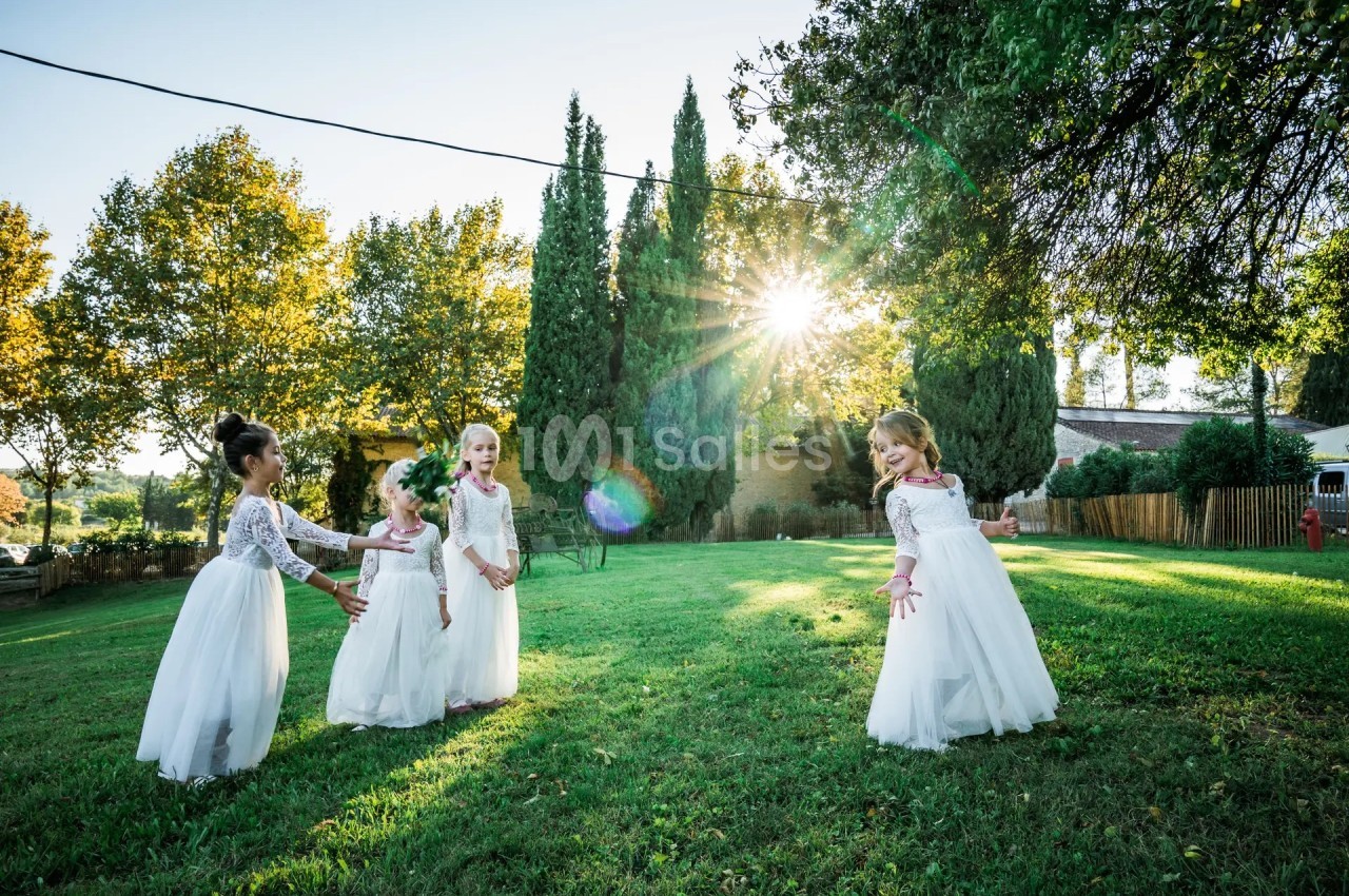 Quatre jeunes filles en robes blanches jouent dans un jardin verdoyant sous un soleil éclatant.
