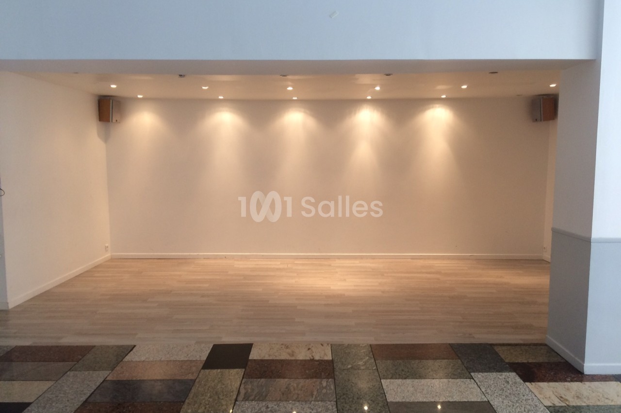 Salle vide avec un mur blanc éclairé par des spots au plafond, parquet clair et sol en carrelage multicolore au premier plan.