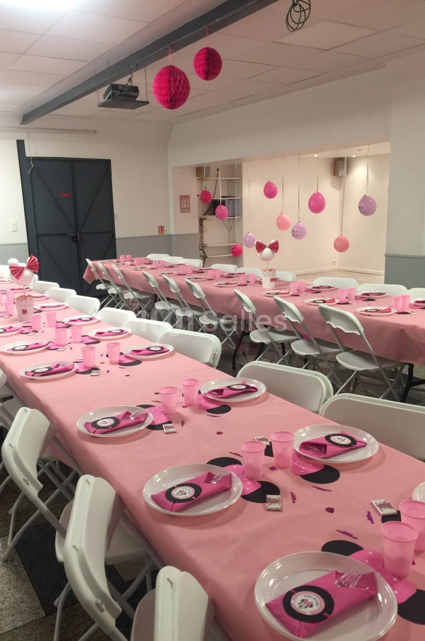 Salle décorée pour une fête avec des tables couvertes de nappes roses, vaisselle assortie et suspensions décoratives.