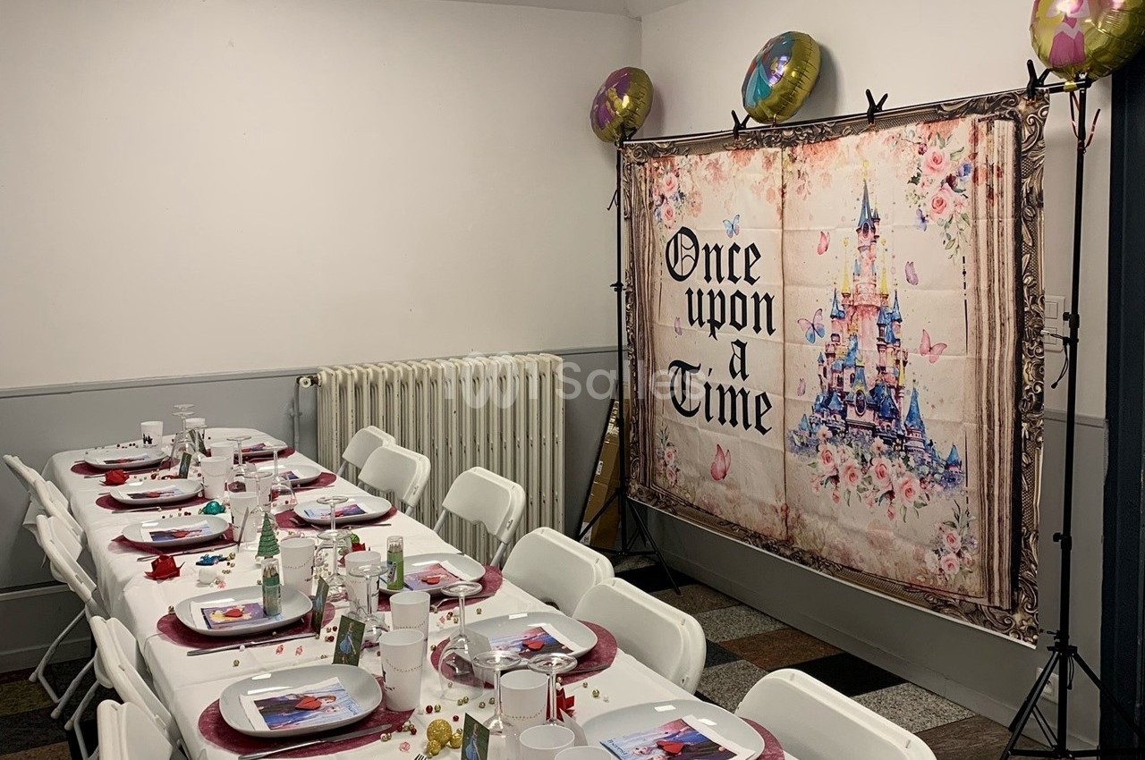 Salle décorée pour une fête avec des tables dressées, un fond illustré d'un château et des ballons colorés.