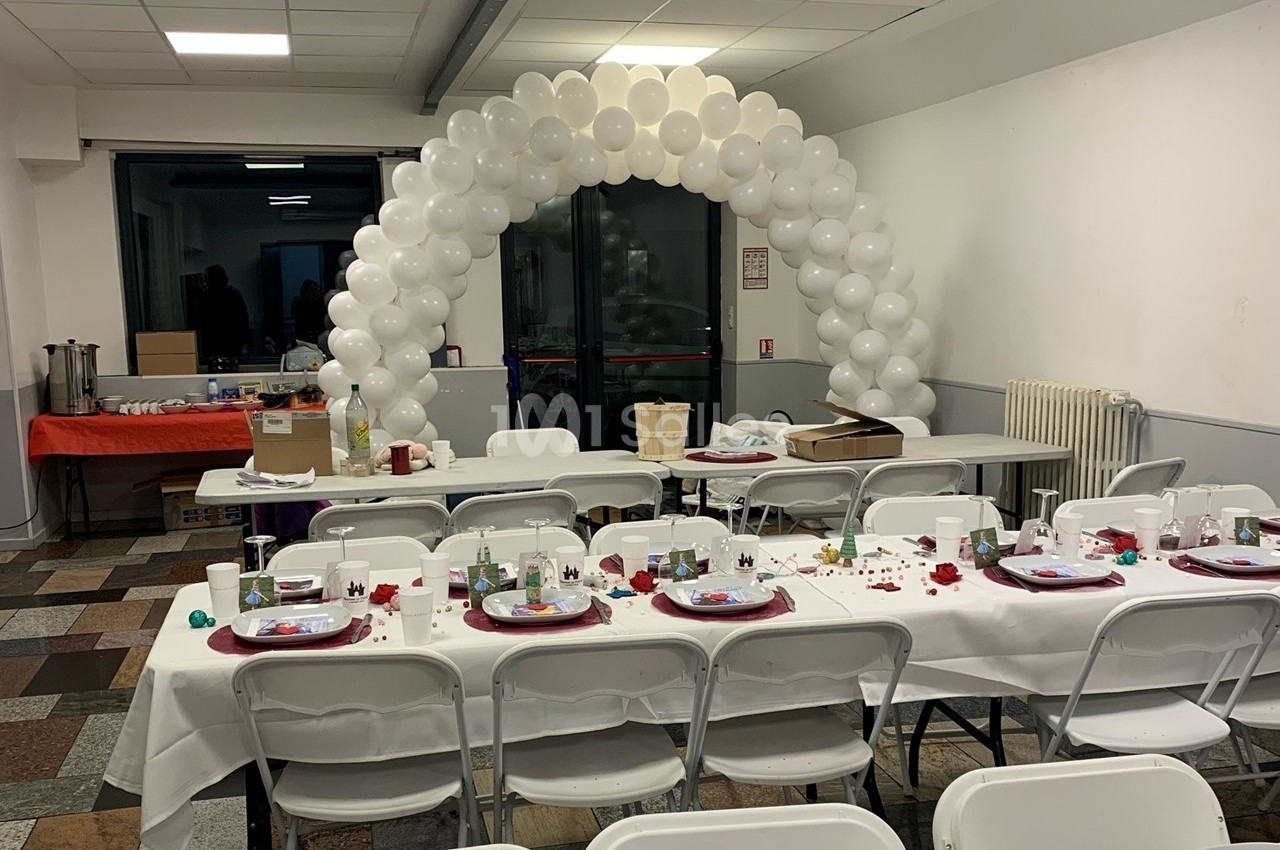 Salle décorée pour un événement avec tables dressées, nappes blanches, et arche de ballons blancs en arrière-plan.