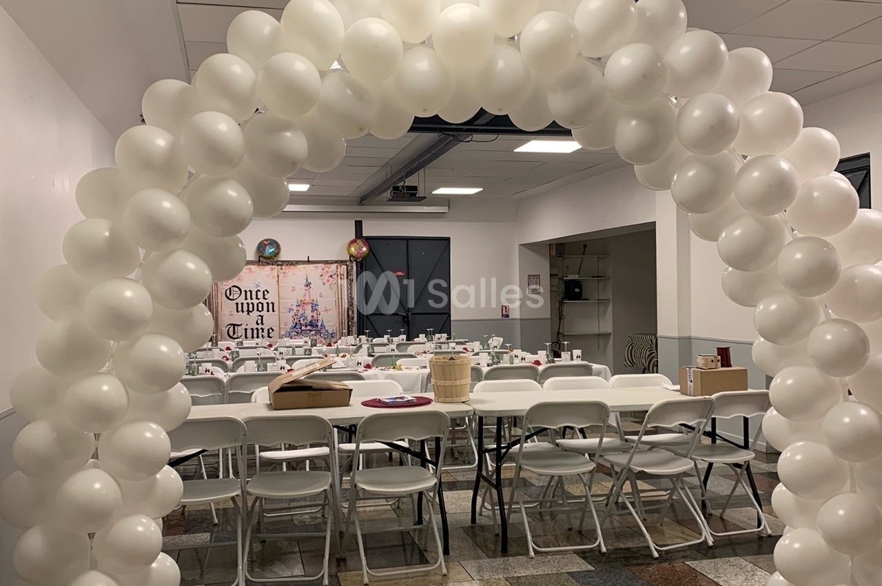 Arche de ballons blancs décorant une salle avec tables et chaises alignées pour un événement festif.