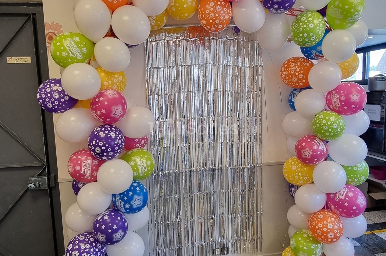 Arche décorative en ballons colorés entourant un rideau métallique argenté dans un espace intérieur.