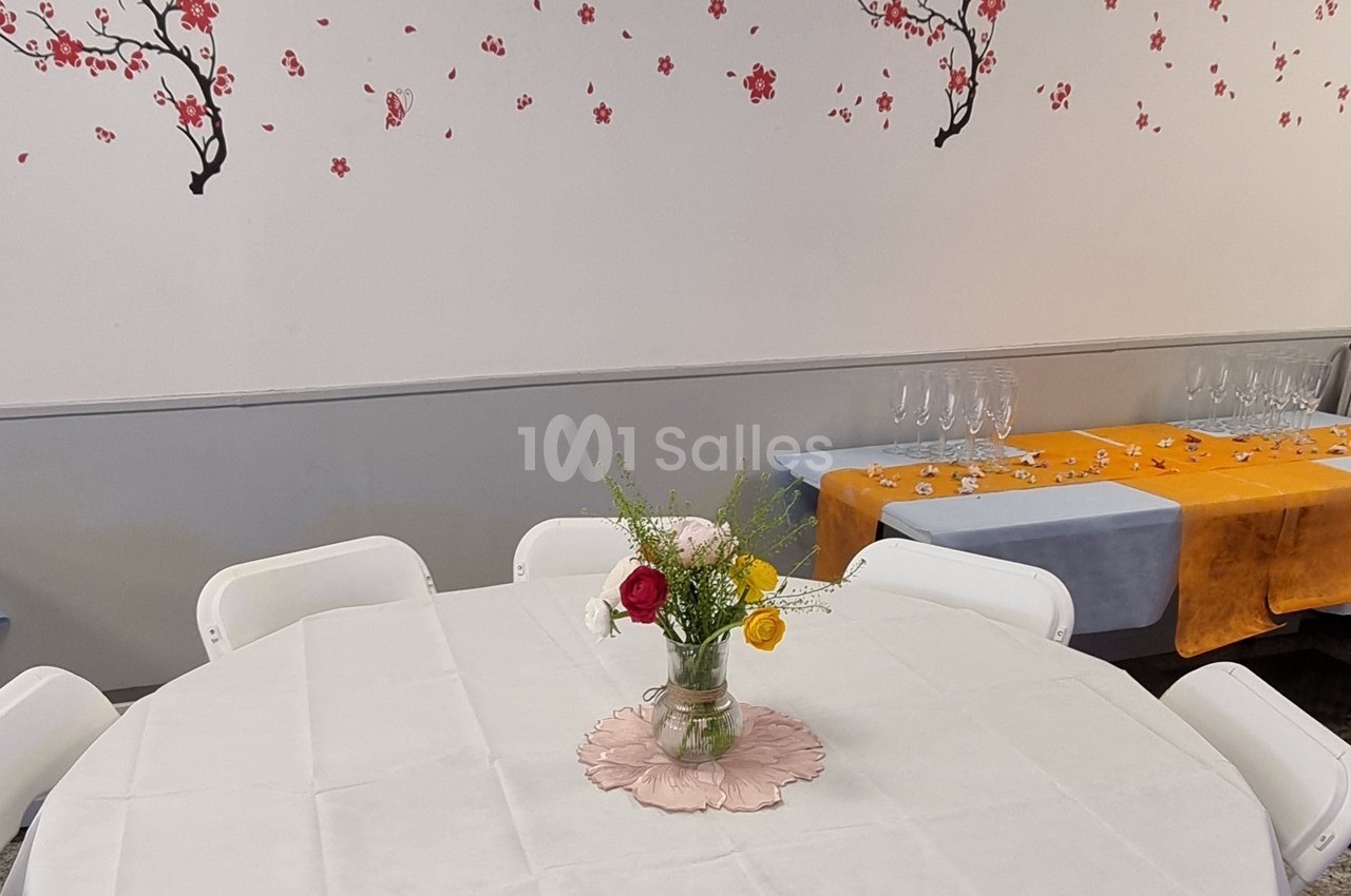 Table ronde dressée avec une nappe blanche, décorée d'un vase de fleurs, devant un mur orné de motifs floraux.