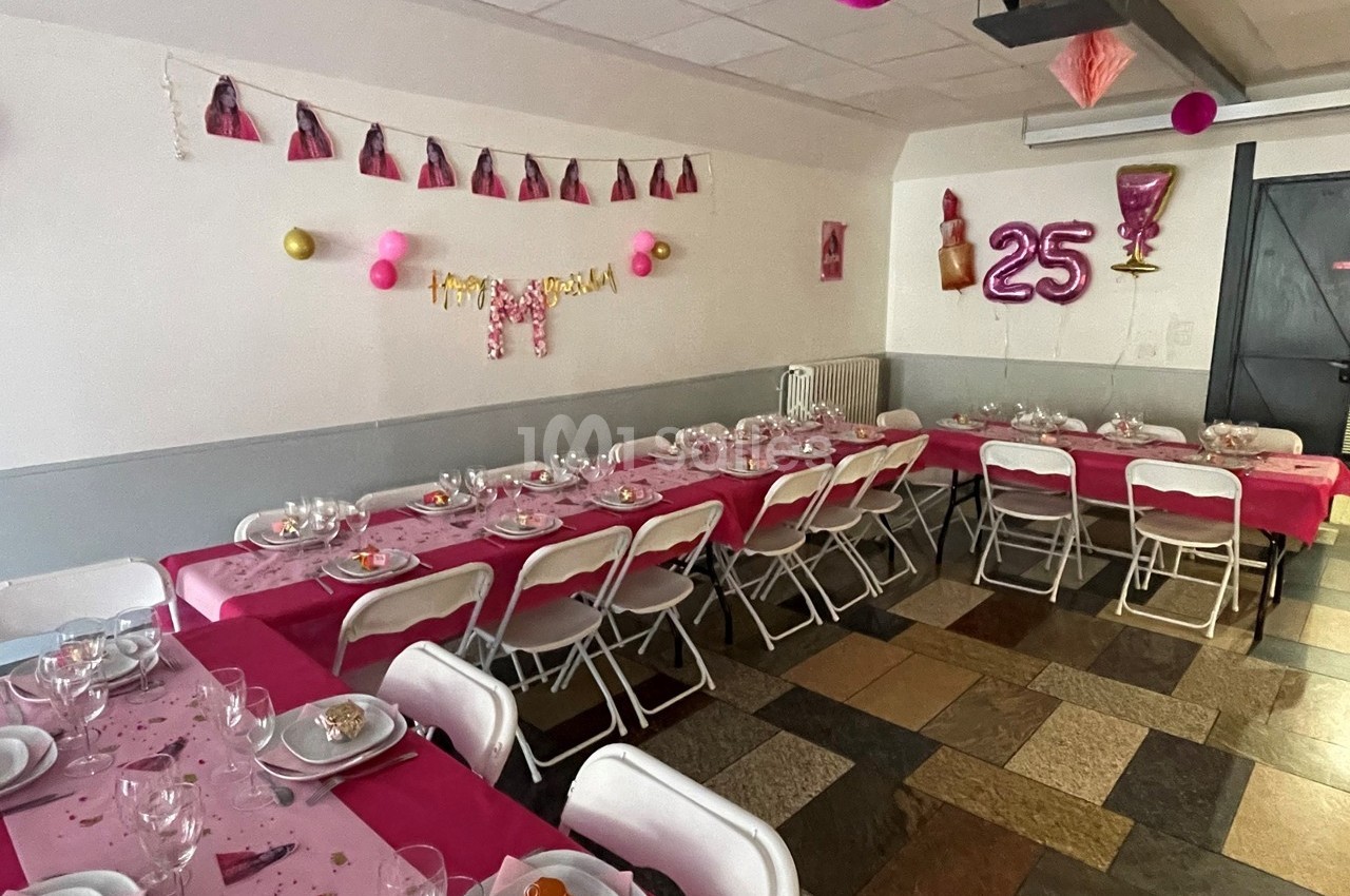 Salle décorée pour un 25e anniversaire avec tables dressées, nappes roses et guirlandes festives accrochées au mur.
