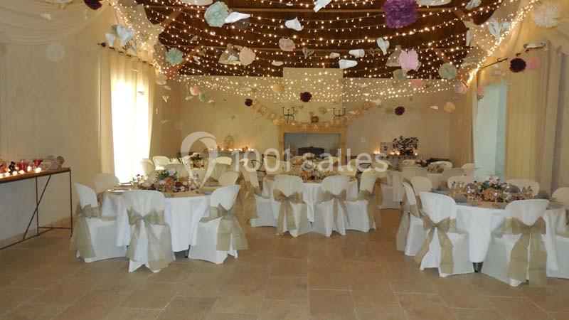 Salle de réception décorée avec des guirlandes lumineuses, des tables rondes dressées et des chaises ornées de nœuds beiges.