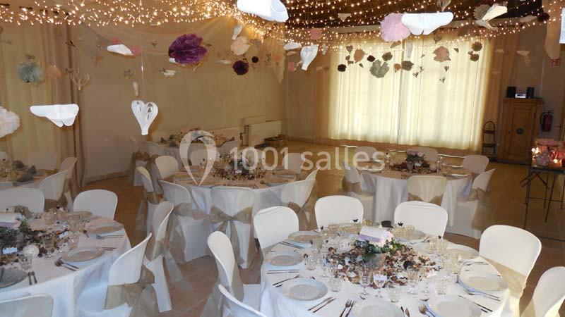 Salle de réception décorée pour un événement, avec tables rondes dressées, chaises blanches et guirlandes lumineuses…