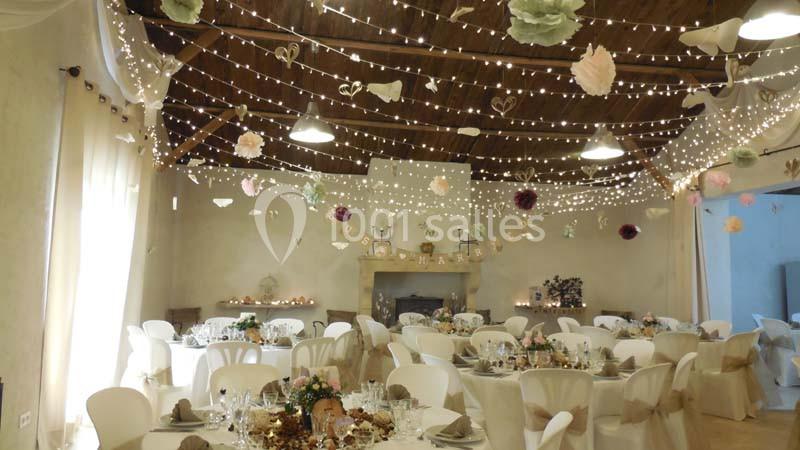 Salle de réception décorée pour un mariage, avec guirlandes lumineuses, tables dressées et ornements suspendus.