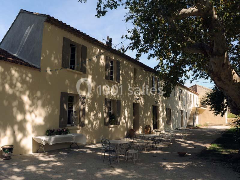 Façade d'une maison provençale avec terrasse ombragée, tables en fer forgé et grand arbre au premier plan.