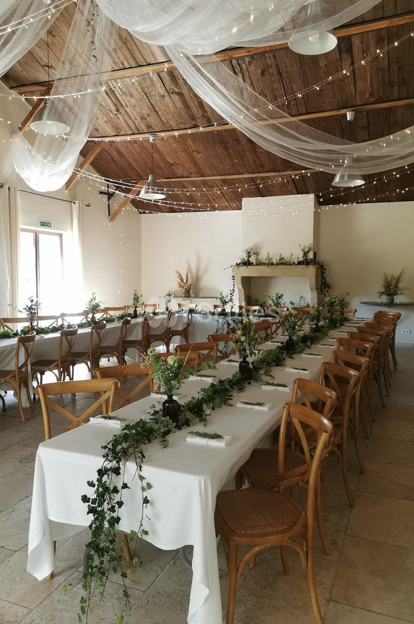 Salle de réception décorée avec des tables alignées, nappes blanches, guirlandes végétales et voilages suspendus.