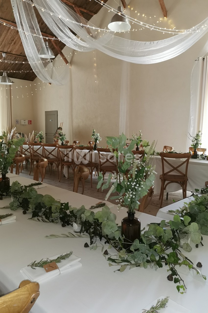 Salle décorée pour un événement avec tables blanches, chaises en bois, guirlandes lumineuses et compositions végétales.