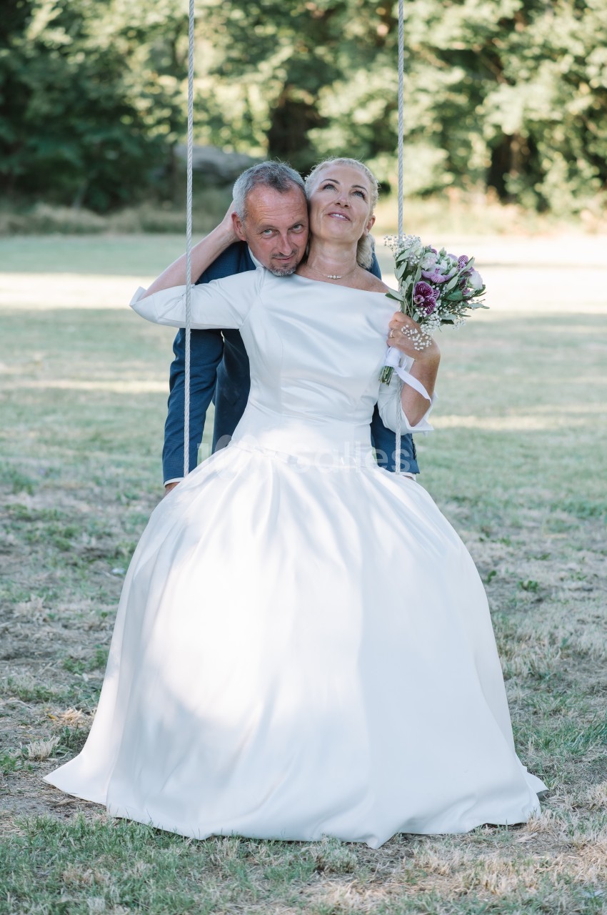 Un couple de mariés souriants, la mariée tenant un bouquet, assis sur une balançoire dans un parc verdoyant.