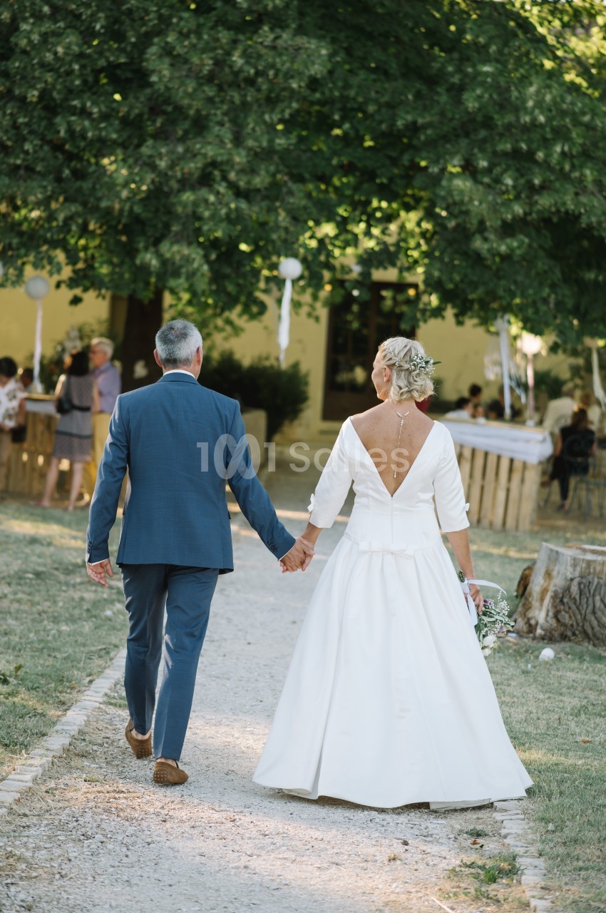 Un couple en tenue de mariage marche main dans la main sur un chemin bordé d'herbe, devant des invités en arrière-plan.