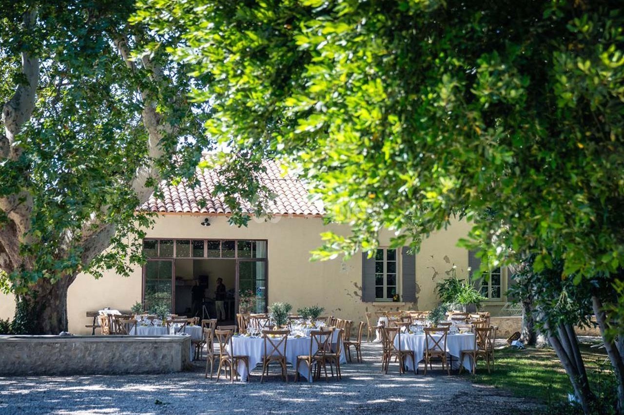 Tables dressées en extérieur sous des arbres, devant une maison aux murs clairs avec des fenêtres à volets verts.