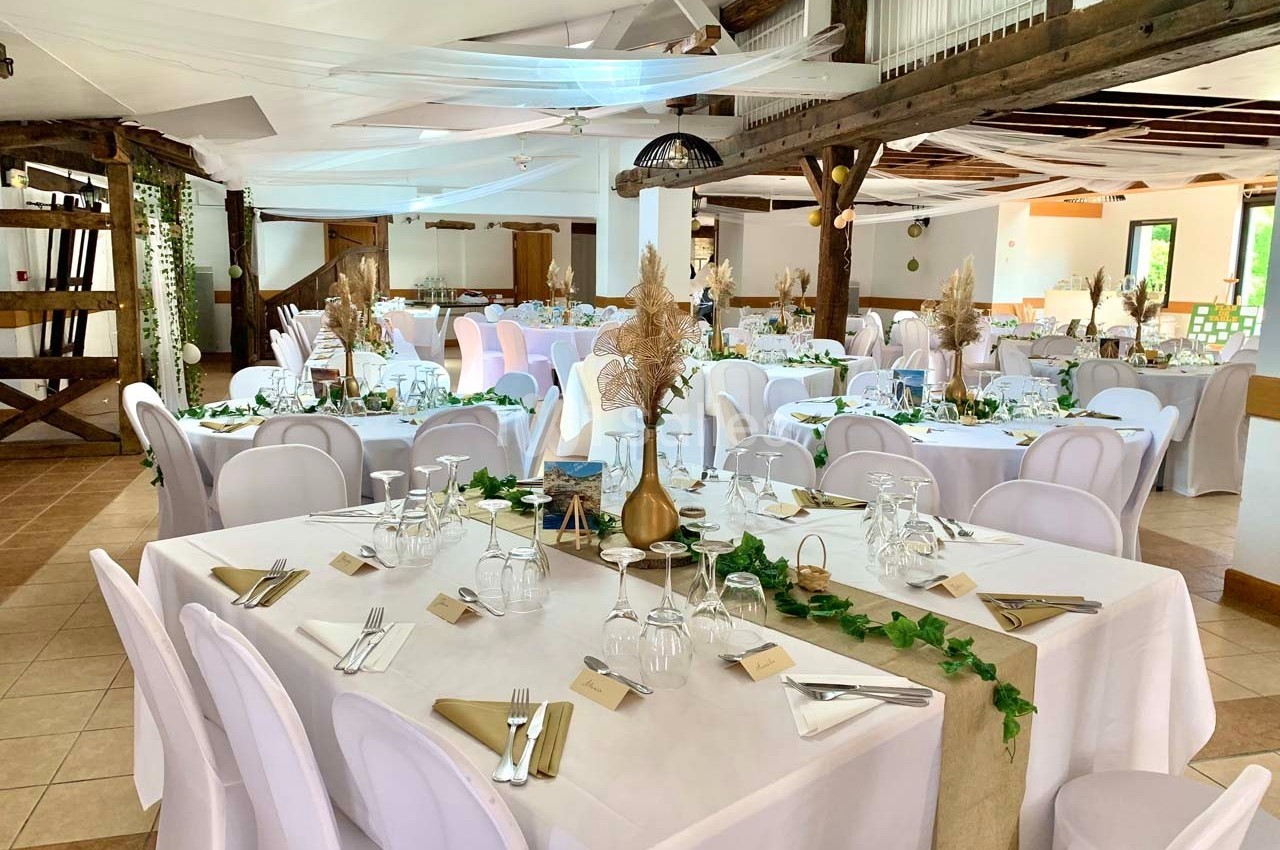 Salle de réception décorée avec des tables dressées, nappes blanches, chaises assorties et ornements végétaux.