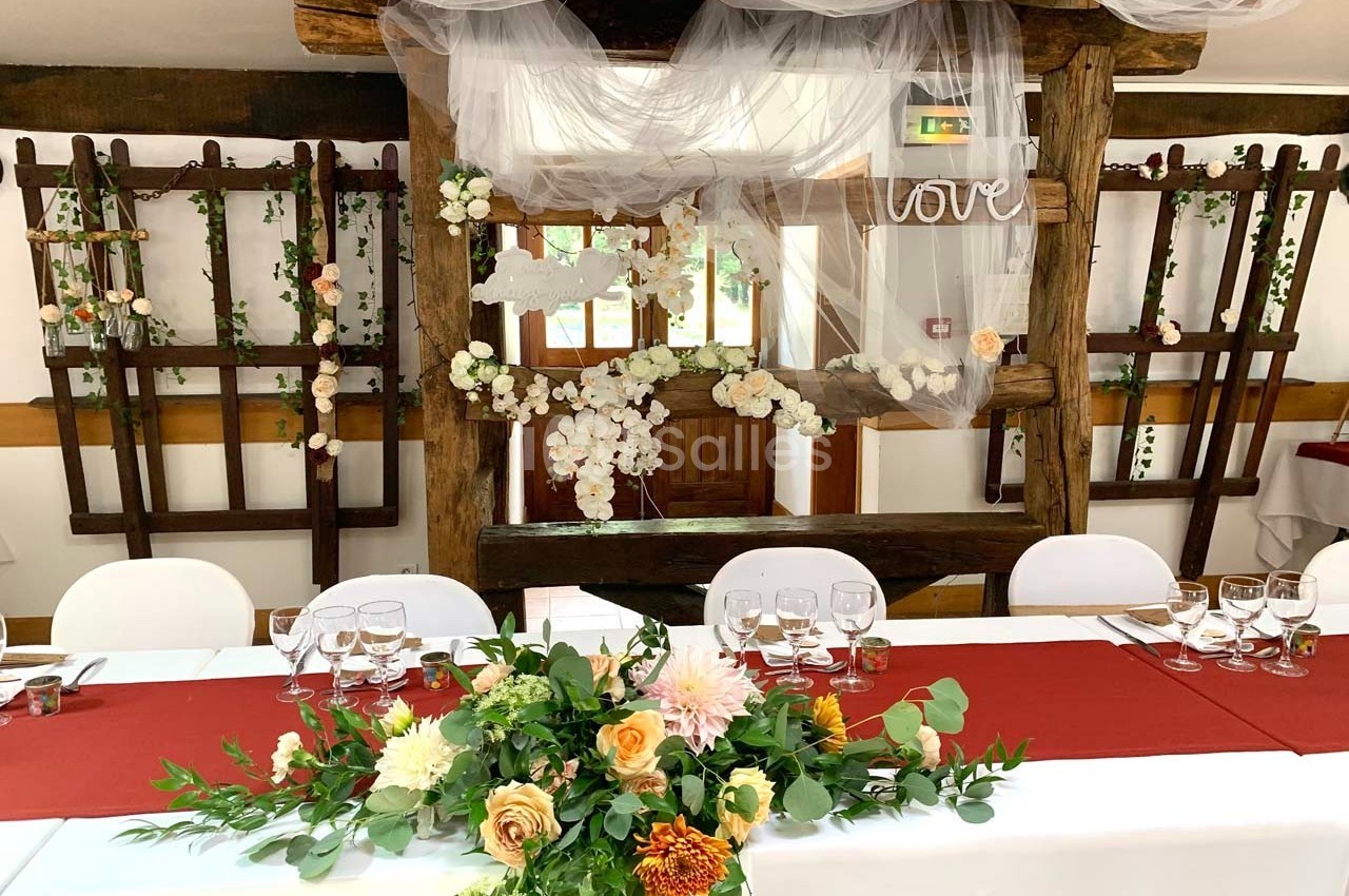 Décoration de salle de réception avec table ornée de fleurs, nappes rouges et arrière-plan en bois décoré de voilages.