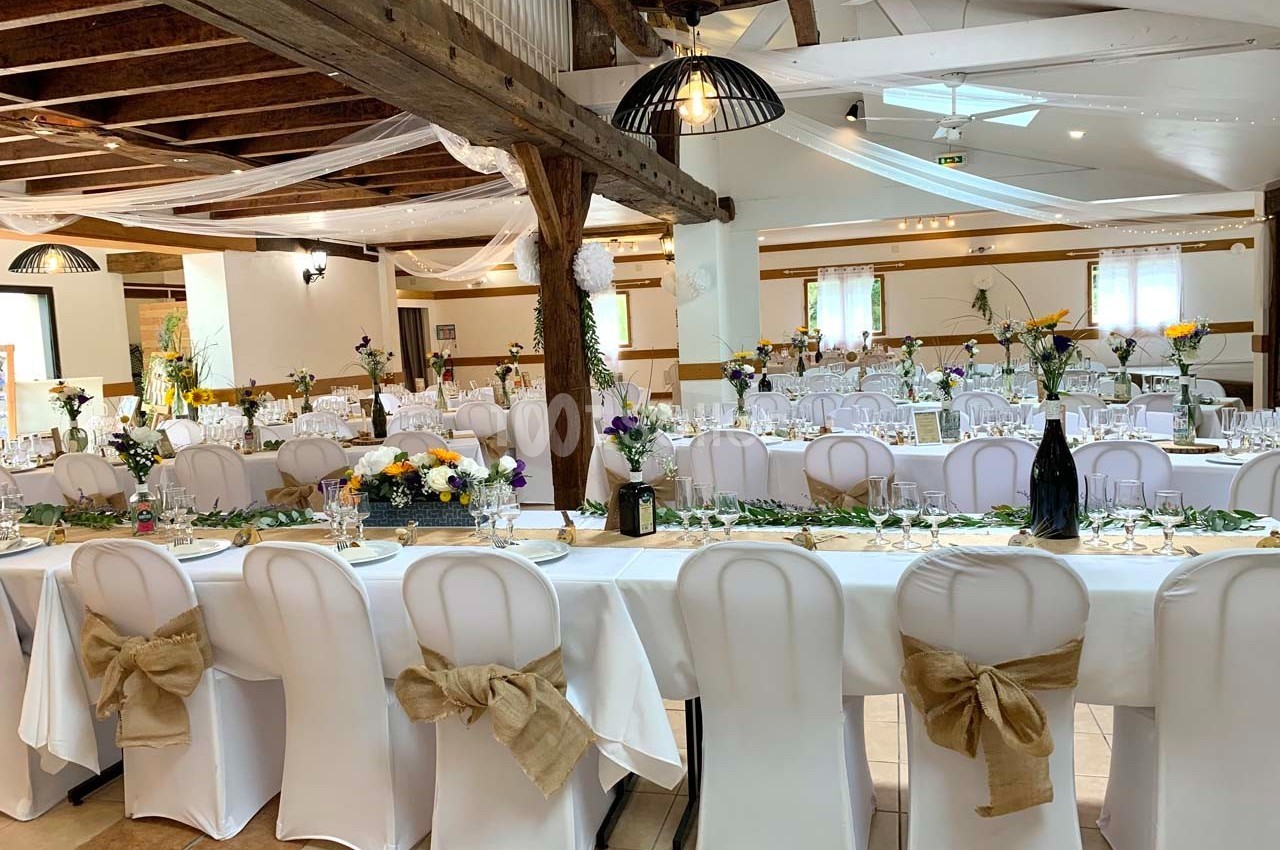 Salle de réception décorée avec des tables dressées, chaises blanches ornées de nœuds en toile de jute et fleurs colorées.