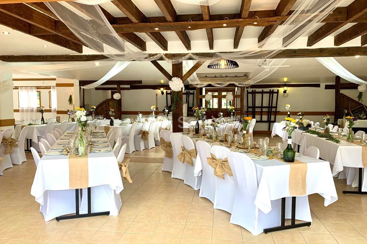 Salle de réception décorée avec des tables dressées, nappes blanches, chaises ornées de nœuds et poutres apparentes.
