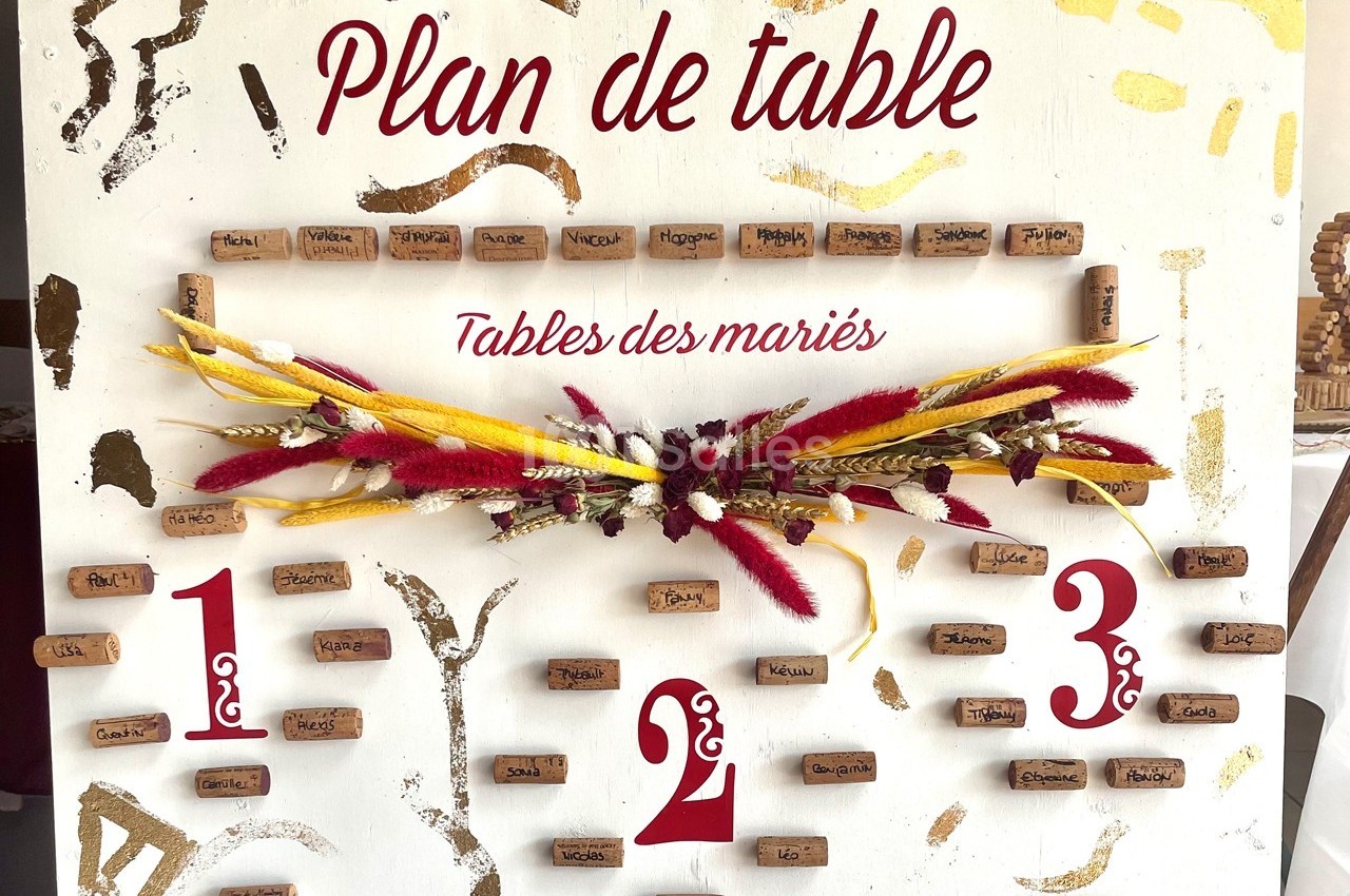 Plan de table décoratif avec des noms inscrits sur des bouchons de liège, orné de fleurs séchées rouges et jaunes.
