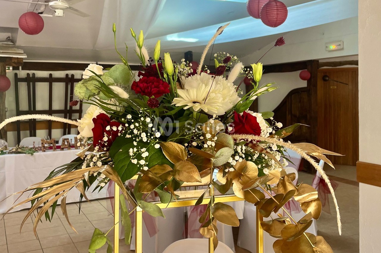 Arrangement floral avec roses rouges, fleurs blanches, gypsophile et feuilles dorées sur un support doré dans une salle…